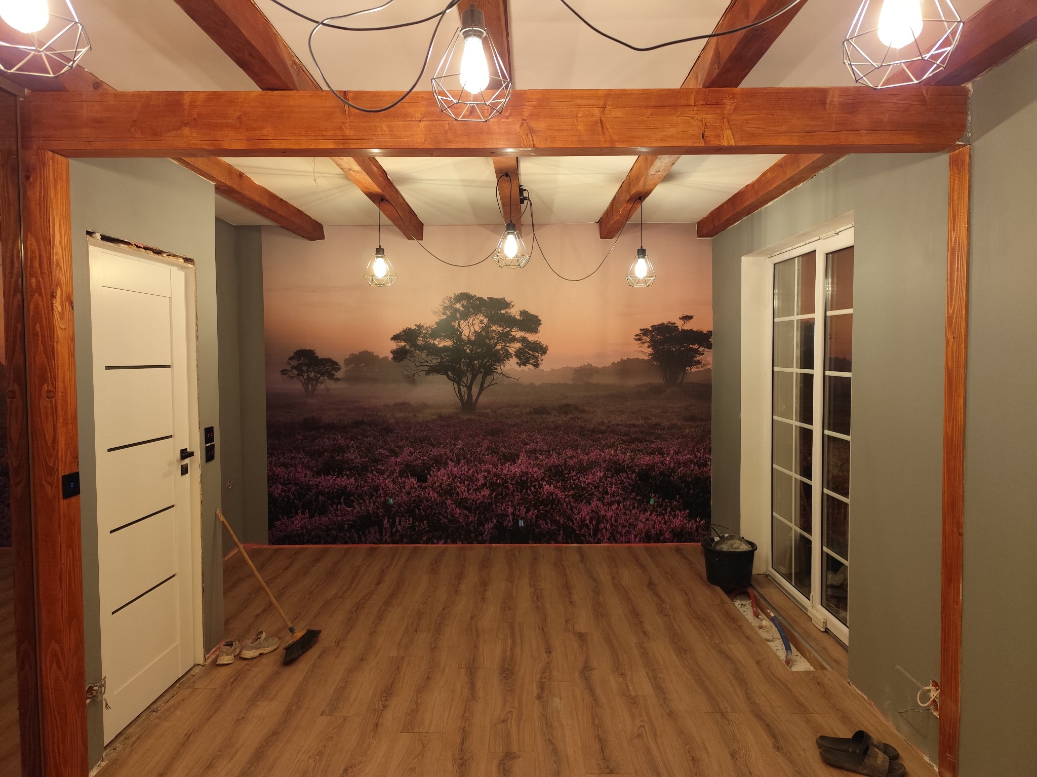 Wnętrze po remoncie: drewniane belki stropowe i podłoga, ściany w szarości, fototapeta z motywem wrzosowiska. Widoczne niedokończone prace przy ościeżnicy i listwach przypodłogowych.