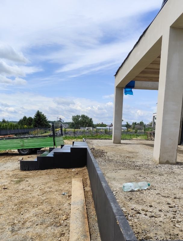 Budowa schodów zewnętrznych z czarnej kostki przy nowoczesnym domu. Widoczny fragment ściany domu i przyczepka. Prace w toku, teren przygotowany pod dalsze etapy.