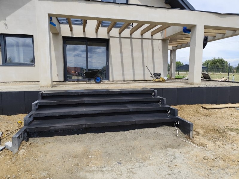 Nowoczesne, czarne schody z kostki brukowej prowadzące do domu z tarasem i pergolą. Widoczna ubijarka i taczka. Prace wykończeniowe w toku.