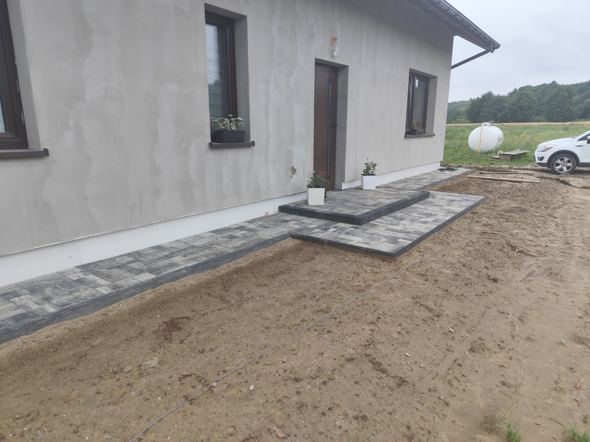Ułożona kostka brukowa prowadząca do drzwi domu, z widocznymi schodami i częściowo przygotowanym terenem. W tle biały samochód i zbiornik.