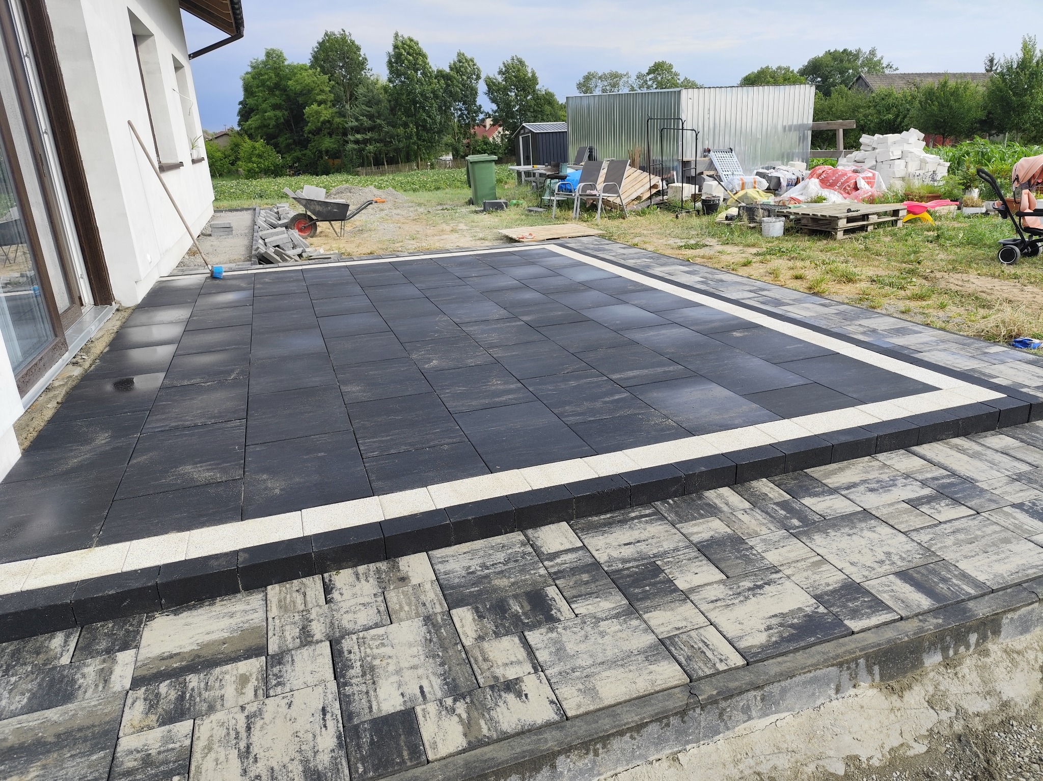 Ułożony taras z ciemnej kostki brukowej z białym obramowaniem, widok na dom i ogród. Uwidocznione detale wykończenia i otoczenie placu budowy.
