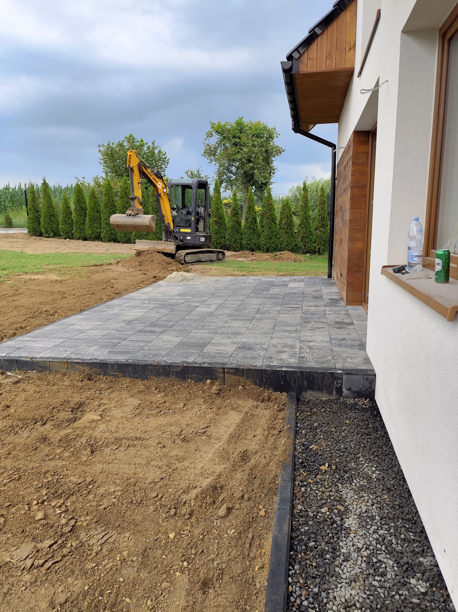 Częściowo ułożona kostka brukowa na posesji w Radgoszczu. Mini koparka w tle przygotowuje teren. Widoczne obrzeża i podsypka.