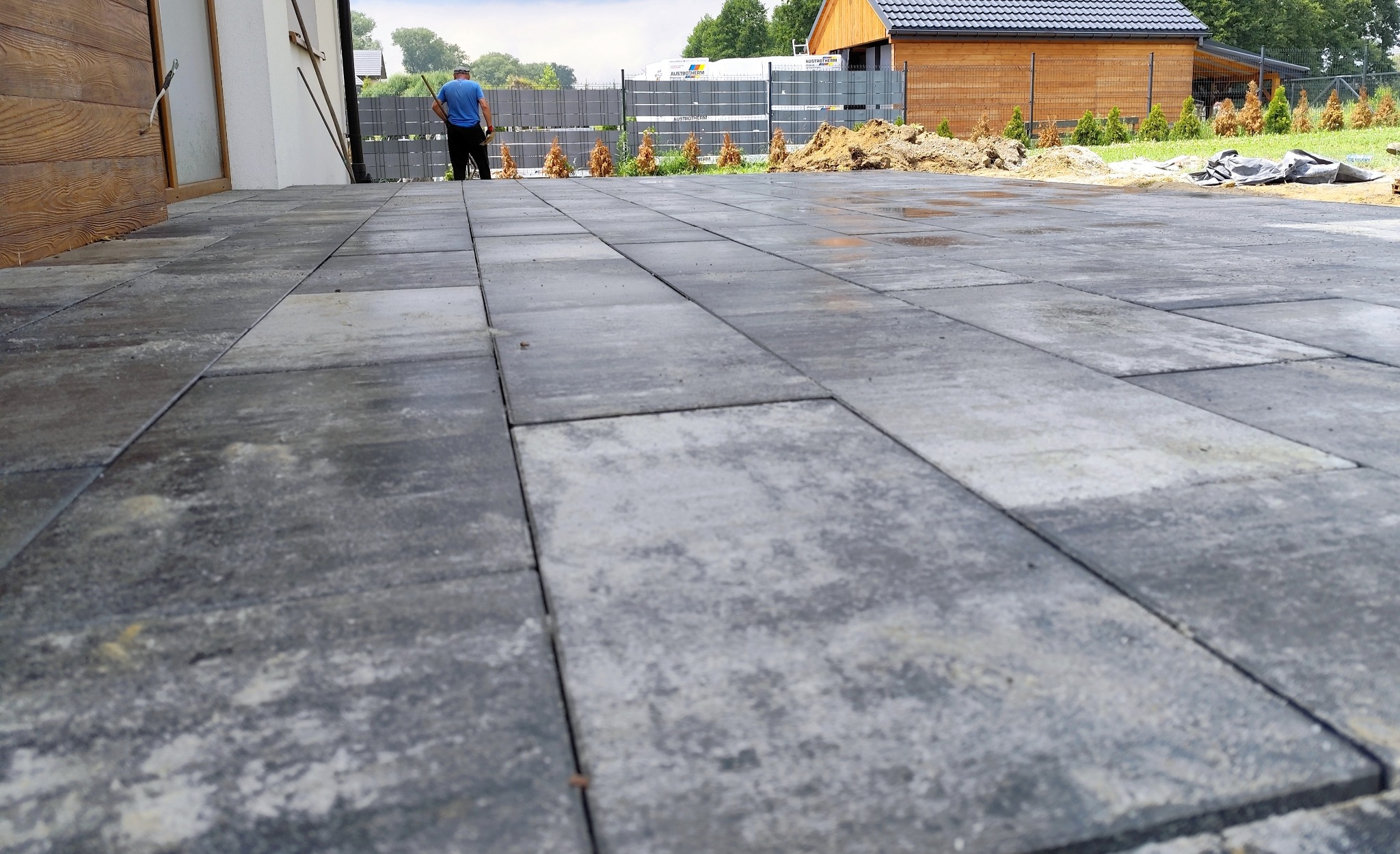 Szara kostka brukowa ułożona na posesji, widoczny fragment domu, ogrodzenie panelowe i sylwetka mężczyzny w oddali. Świeżo ułożona nawierzchnia, praca w toku.