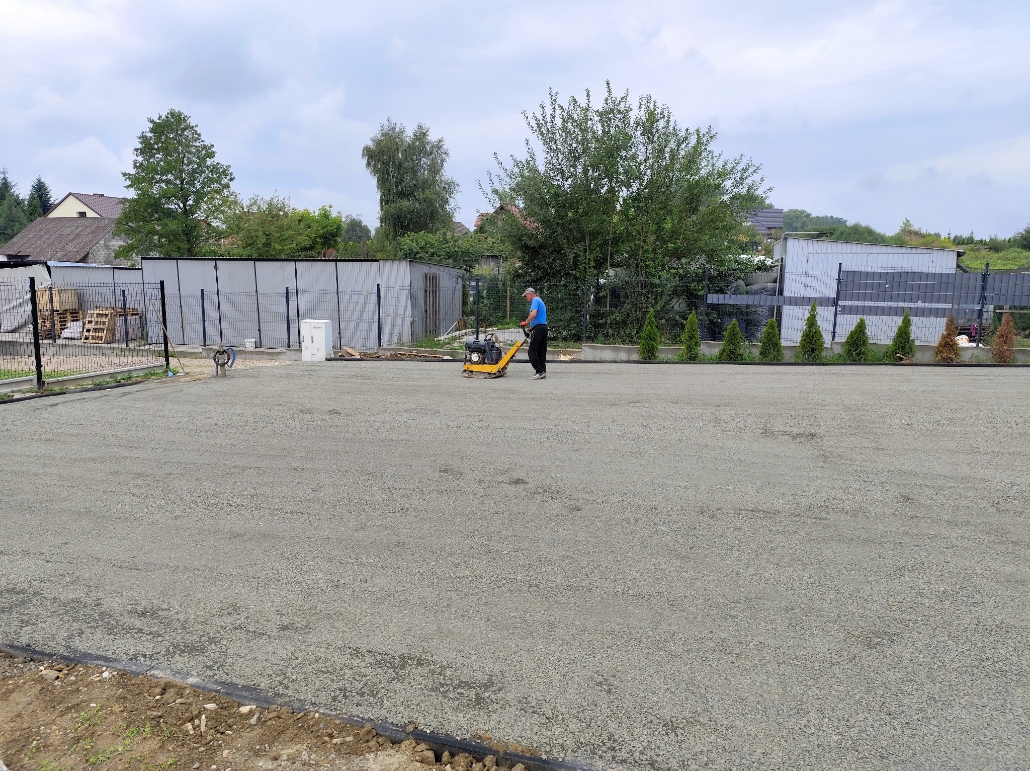 Pracownik utwardza teren podłoża zagęszczarką w otoczeniu zieleni i zabudowań gospodarczych. Widoczna przygotowana, utwardzona powierzchnia z obrzeżami.