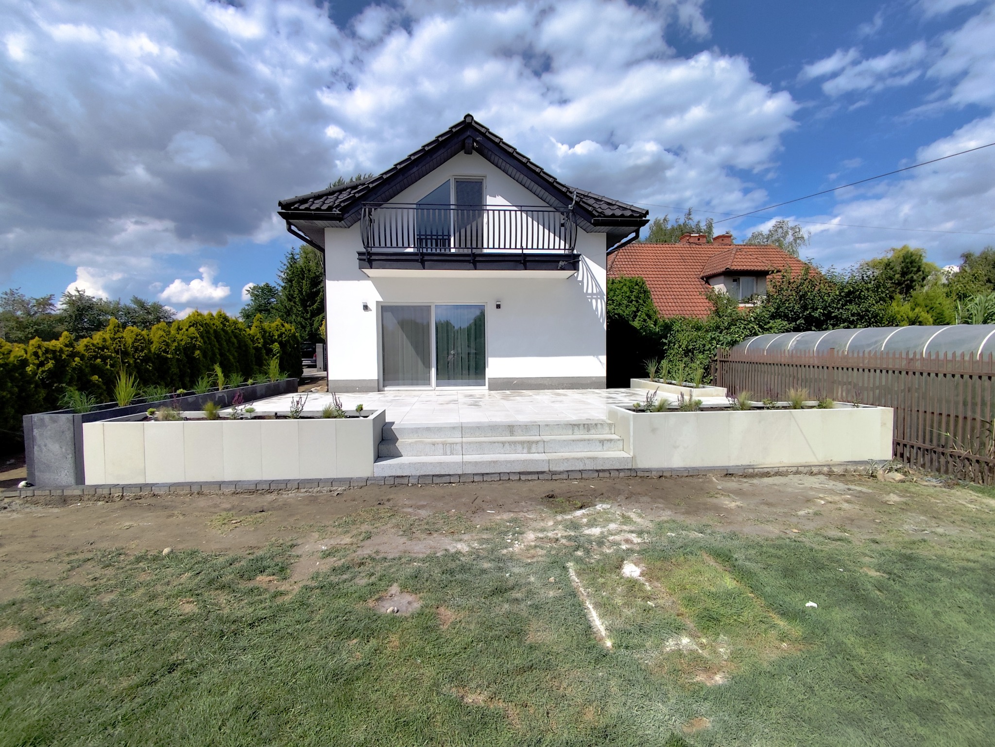 Nowoczesny dom z tarasem wyłożonym jasną kostką, schody z kostki, minimalistyczne rabaty z roślinami. Błękitne niebo z chmurami.