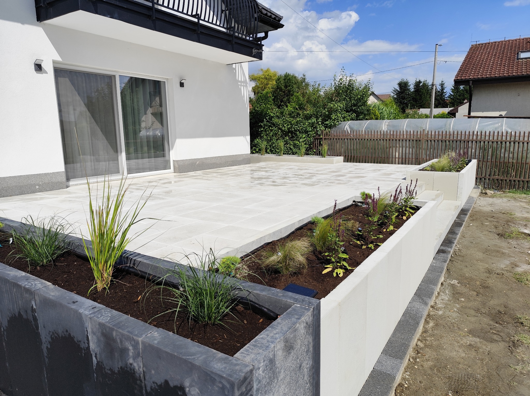 Nowoczesny taras z jasnej kostki brukowej, obrzeża z donicami z roślinami, biały budynek w tle, błękitne niebo. Układ kostki w regularny wzór, minimalistyczny design.