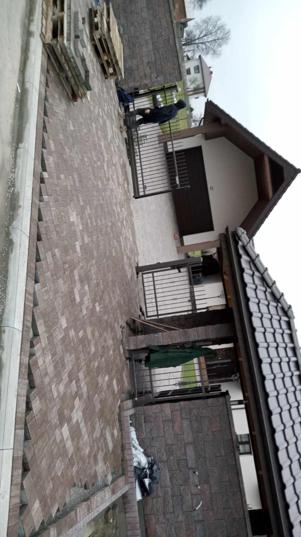 Ułożona kostka brukowa w jodełkę w odcieniach brązu, obok posesji z bramą i fragmentem budynku z ciemnym dachem. Na paletach ułożona kostka. Widok z góry.