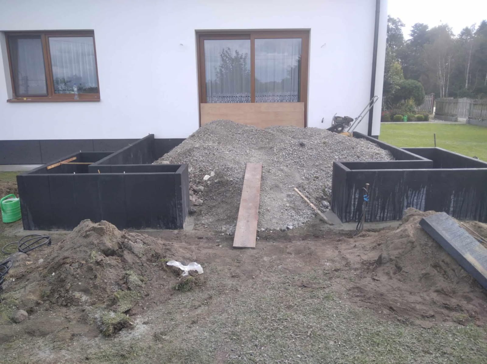 Fundamenty domu w trakcie budowy: czarne bloczki fundamentowe, góra żwiru, narzędzia budowlane, widok na biały budynek i trawnik. Etap prac fundamentowych.
