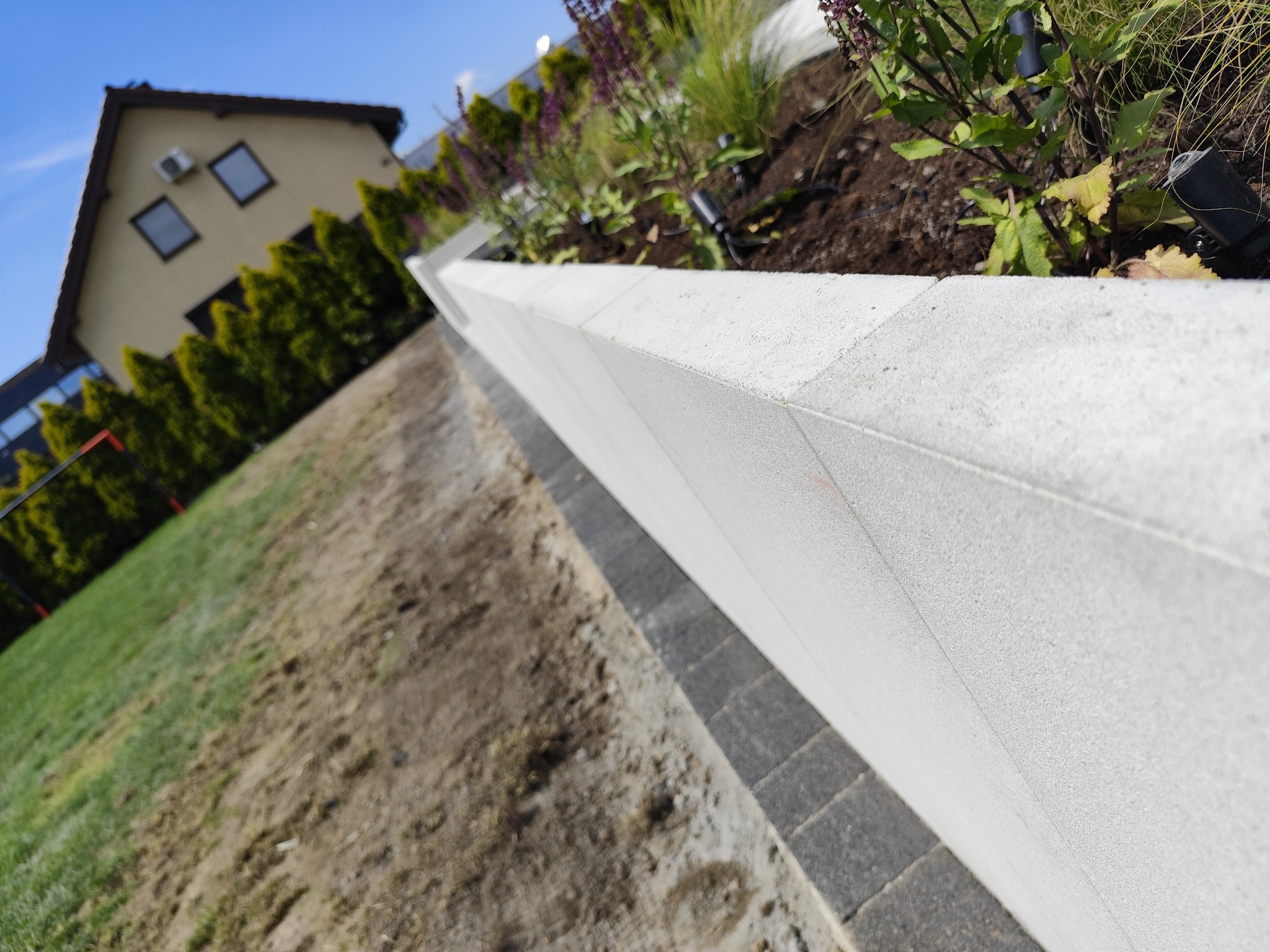 Nowoczesny murek oporowy z jasnych bloczków betonowych, oddzielający trawnik od rabaty z roślinami. W tle dom i rząd tuj.