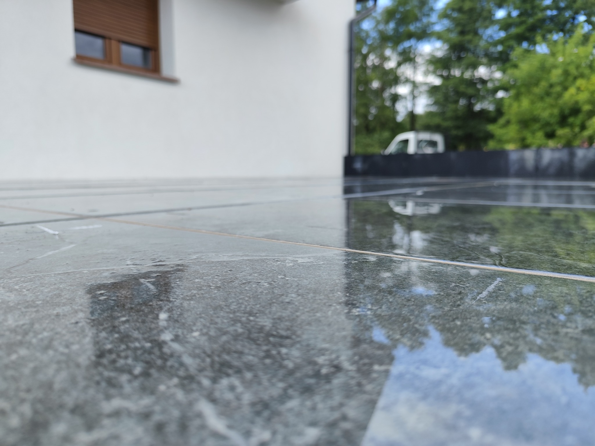 Połyskująca, ciemnoszara kostka brukowa na tarasie domu z białymi ścianami i oknem z brązowymi roletami. W tle zieleń drzew i częściowo widoczny samochód.