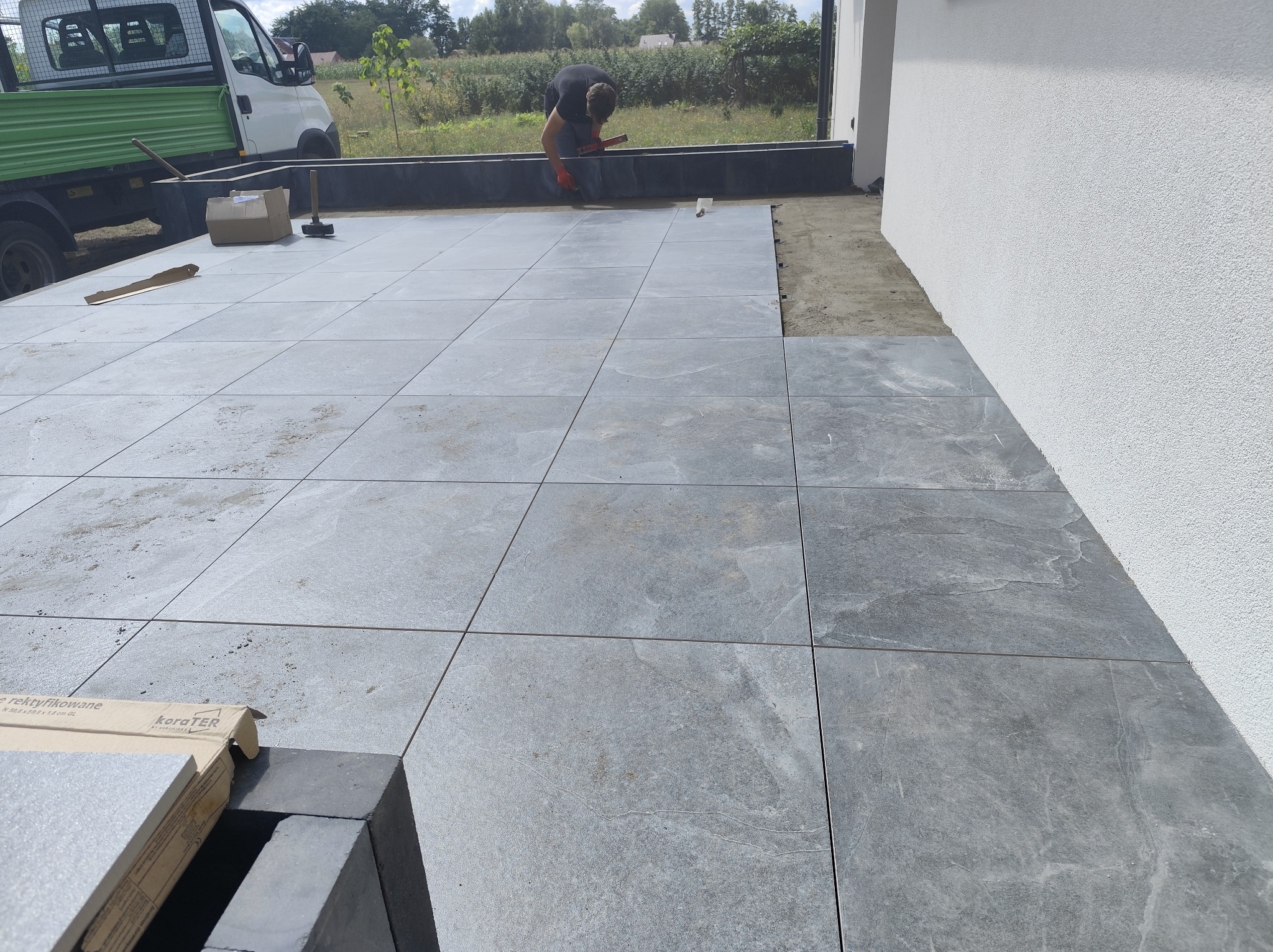 Układanie płyt tarasowych z szarego kamienia na zewnątrz domu. Widoczny fragment samochodu dostawczego i pracownik w trakcie montażu. Część płyt ułożona, część jeszcze nie.
