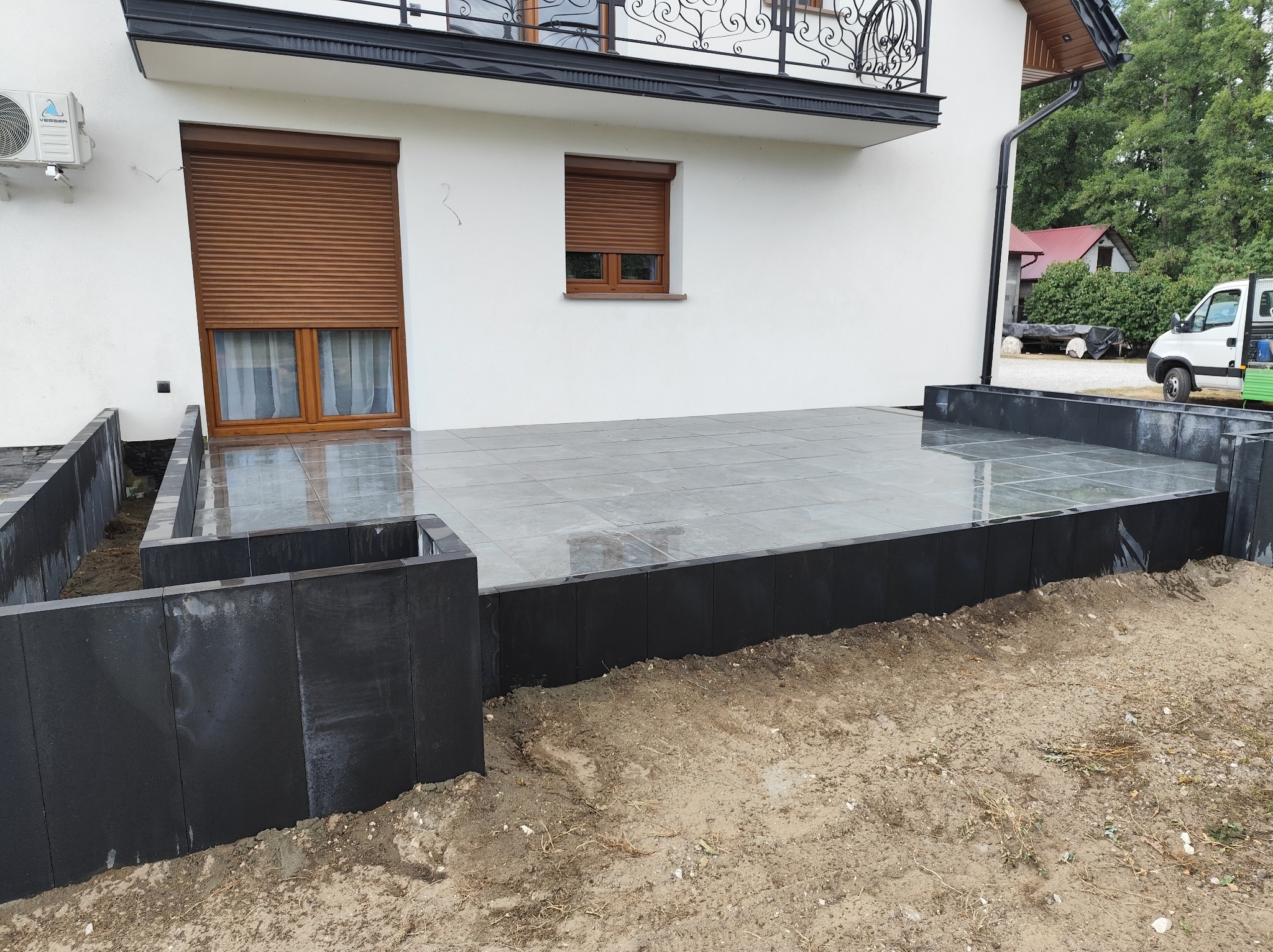 Nowoczesny taras z szarych płyt, obramowany czarnymi elementami, przylegający do białego budynku z brązowymi roletami. Widoczny fragment ogrodu i zaparkowany samochód.