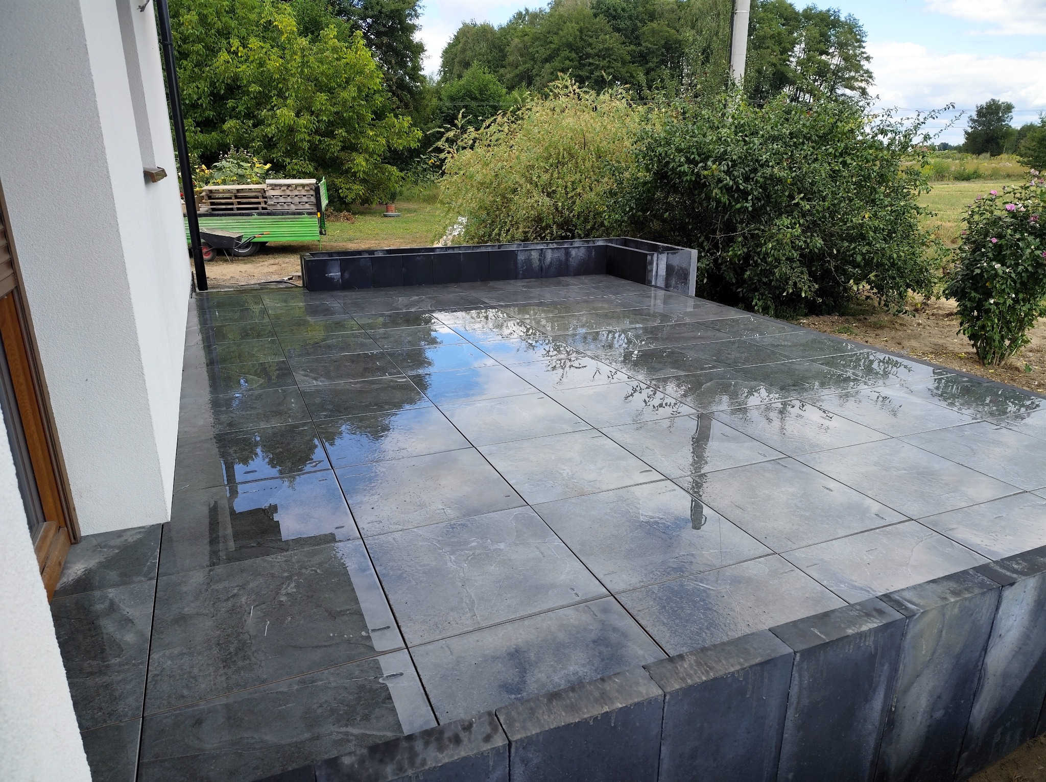 Świeżo ułożony, grafitowy taras z kostki brukowej po deszczu, z widocznym odbiciem nieba. W tle zieleń ogrodu i fragment ściany domu.