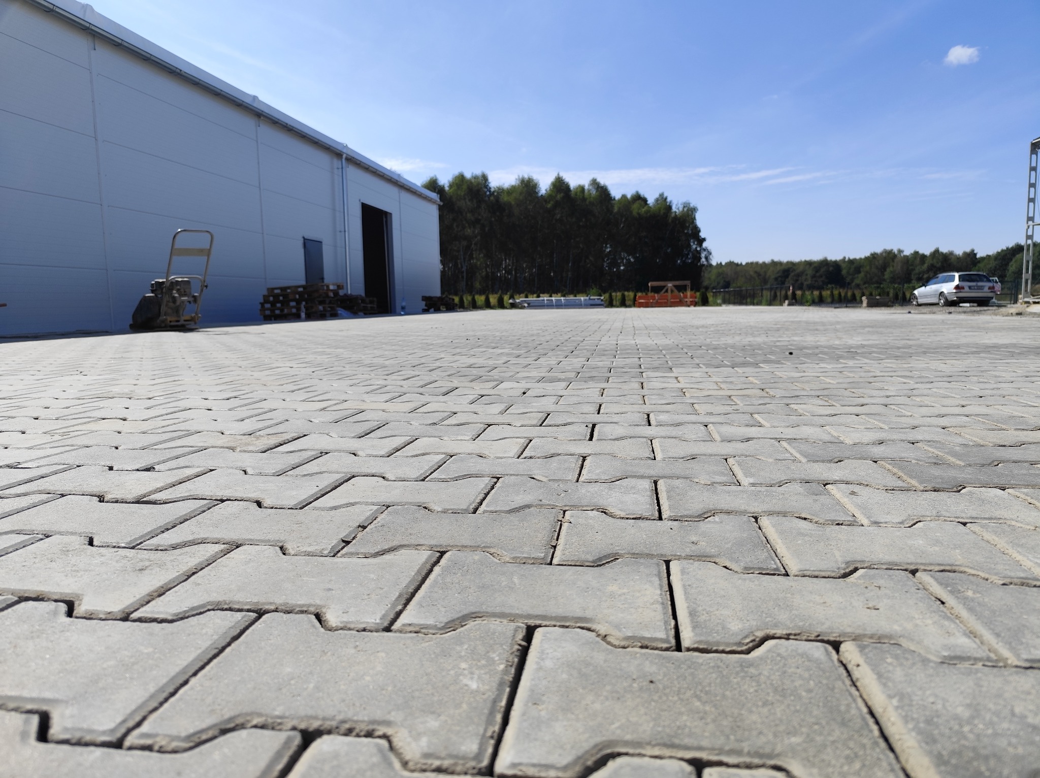 Szara kostka brukowa ułożona na dużym placu przed magazynem, z ubijarką w tle. Perspektywa z dołu podkreśla regularność wzoru. Błękitne niebo i zieleń drzew w oddali.