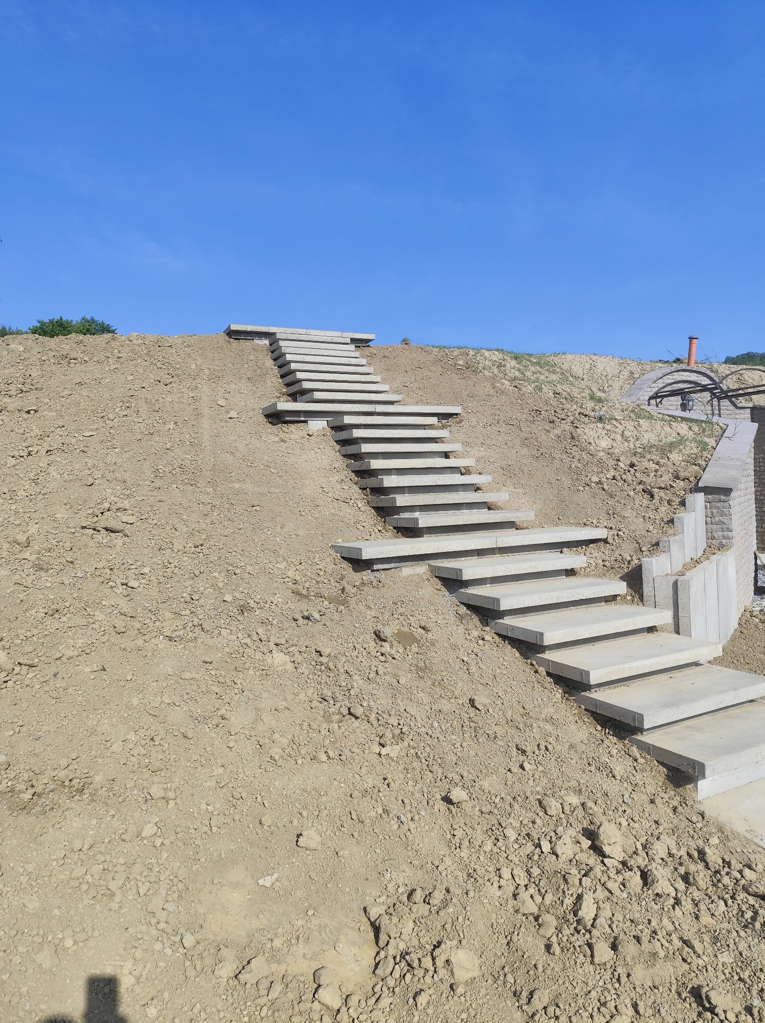 Schody z betonowych płyt prowadzące na wzgórze pod błękitnym niebem. Elementy architektury ogrodowej w tle, ziemia wokół schodów. Ujęcie z dołu.