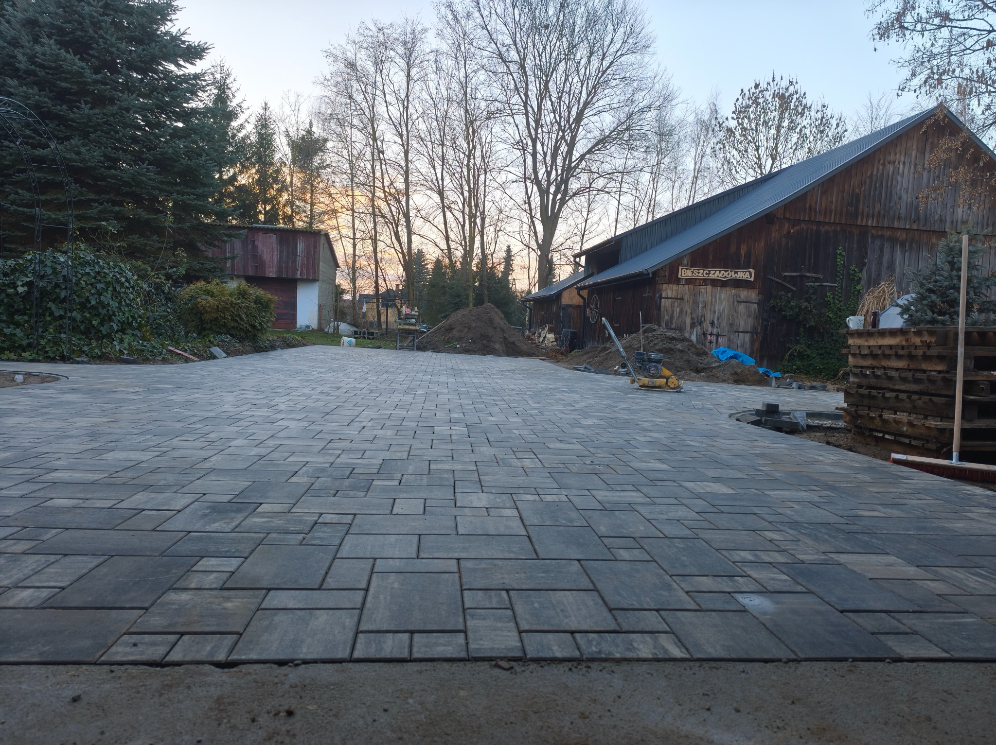 Ułożona kostka brukowa na podjeździe w odcieniach szarości, z widoczną ubijarką i stertą ziemi w tle, przy drewnianym budynku gospodarczym w wiejskim stylu.