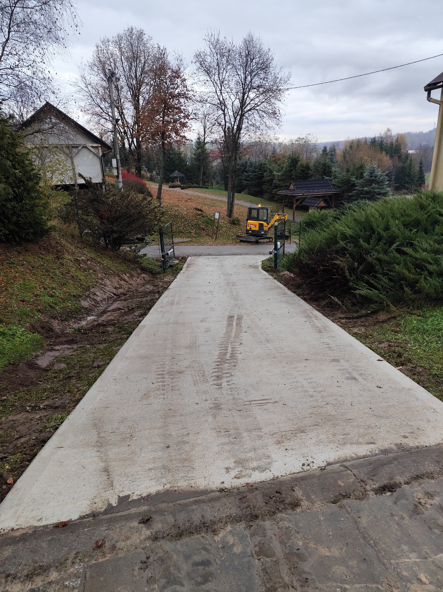 Betonowy podjazd do posesji z widocznym mini-koparką w tle, otoczony zielenią i drzewami w jesiennej scenerii. Ślady opon na świeżym betonie.