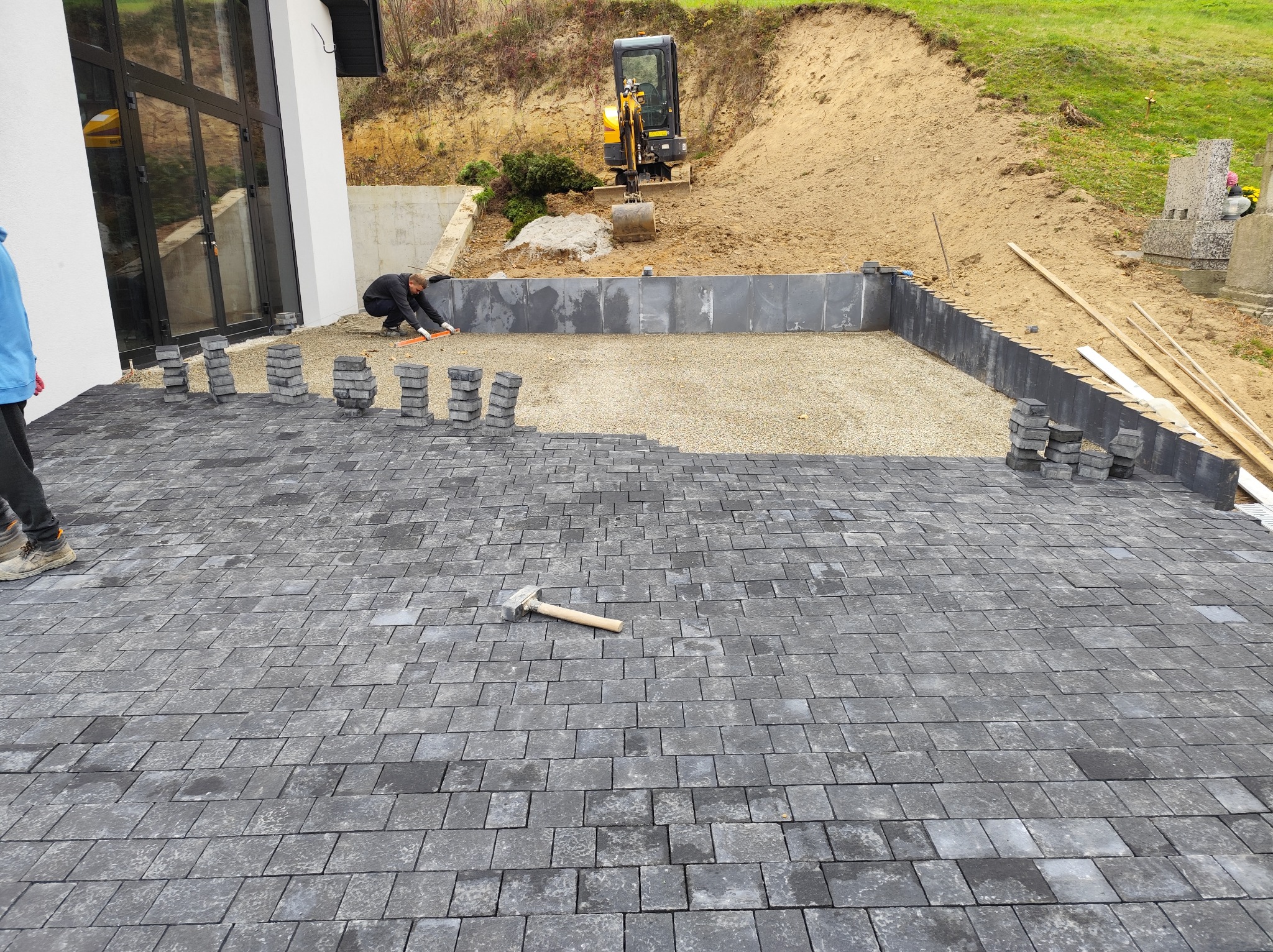 Częściowo ułożona, szara kostka brukowa na posesji z mini koparką w tle. Widoczny pracownik przy układaniu, stosy kostki i młotek brukarski.