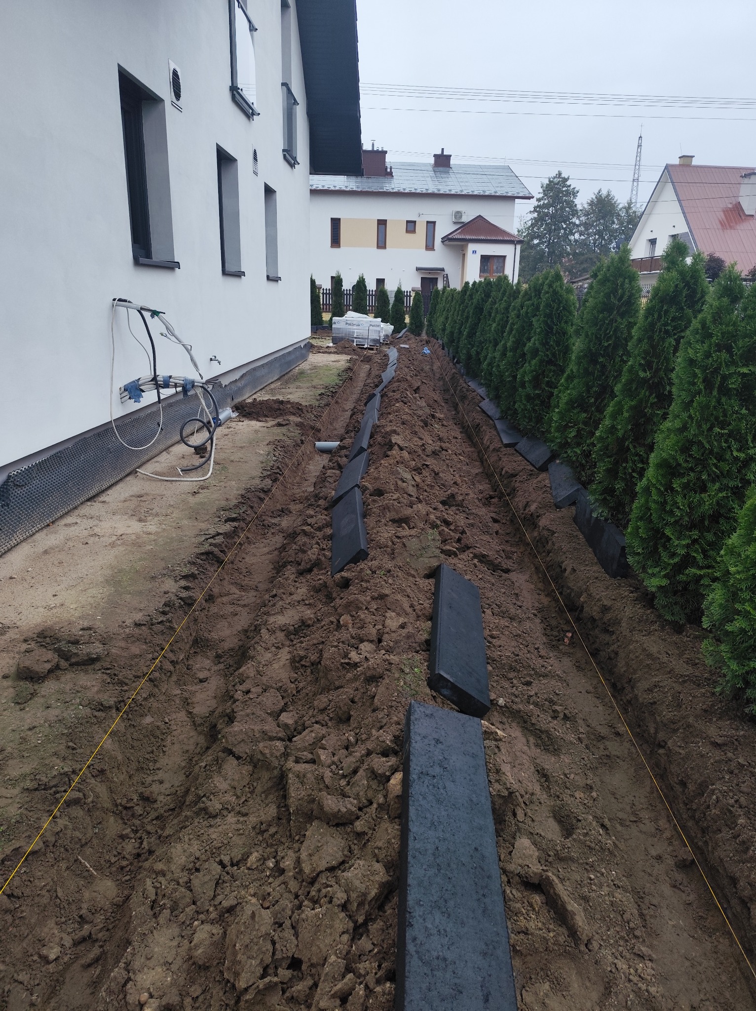 Wykopany rów wzdłuż żywopłotu z czarnymi obrzeżami, przygotowany do montażu. Widoczne rury i przewody przy budynku. Prace ziemne w toku.