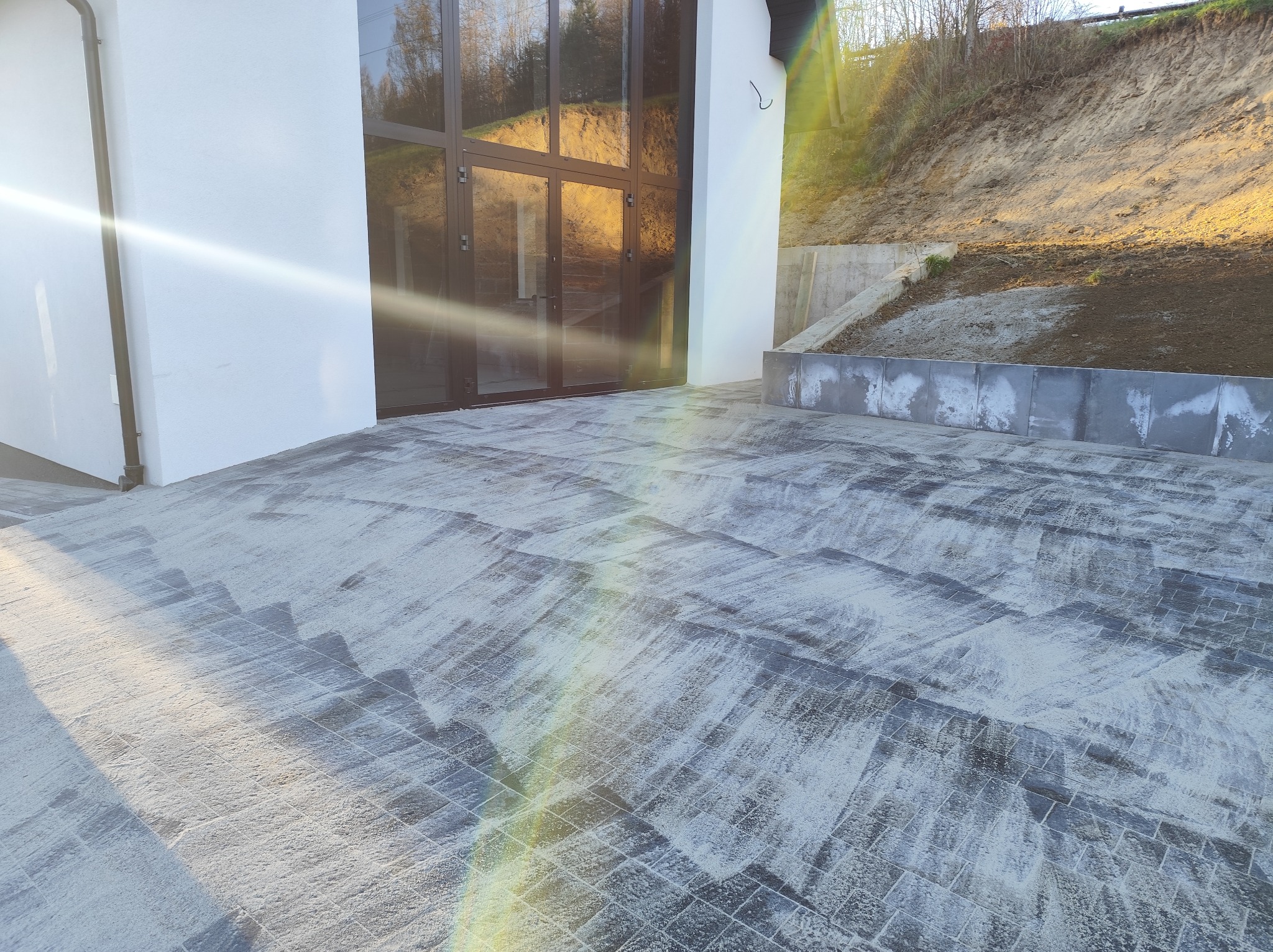 Nowoczesna posesja z szarą kostką brukową ułożoną na podjeździe, widok na przeszklone drzwi garażowe i skarpę z elementami betonowymi w tle, w słoneczny dzień.