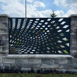 Profesjonalne cięcie CNC - Nowoczesne ogrodzenie z metalu o geometrycznym wzorze, osadzone na kamiennej podmurówce. Widoczne niebo i zieleń w tle.