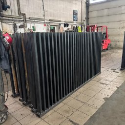 Profesjonalne cięcie CNC - Czarne, metalowe elementy konstrukcyjne z pionowymi prętami, ustawione w hali produkcyjnej obok spawarki i wózka widłowego. Gotowe do montażu.