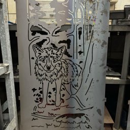 Profesjonalne cięcie CNC - Metalowy panel z wyciętym motywem wilka na tle lasu, wykonany techniką CNC, oparty o metalowy regał. Widoczne ślady obróbki i zabrudzenia.