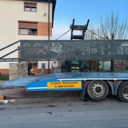 Profesjonalne cięcie CNC - Transport bramy z wycinanymi laserowo wzorami wiejskimi na lawecie pomocy drogowej. W tle dom z oknem, w którym stoi osoba. Widoczny traktor i fragmenty montażu.