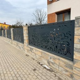 Profesjonalne cięcie CNC - Nowoczesne ogrodzenie panelowe z dekoracyjnym, ażurowym wzorem, osadzone na niskim murku z kamiennych bloczków. Widok na chodnik i domy w tle.