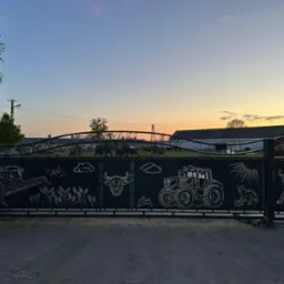 Czarna brama wjazdowa z motywami rolniczymi (traktor, kombajn, zwierzęta) na tle zachodzącego słońca. Metaloplastyka z dbałością o detal.