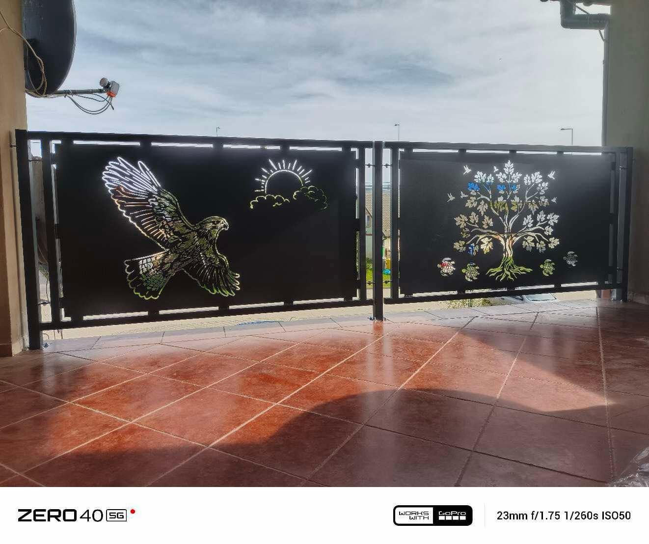 Metalowa brama wjazdowa z dekoracyjnymi wycięciami: orzeł w locie i drzewo. Ciemne tło bramy kontrastuje z jasnym tłem nieba. Widoczne płytki na podłodze.