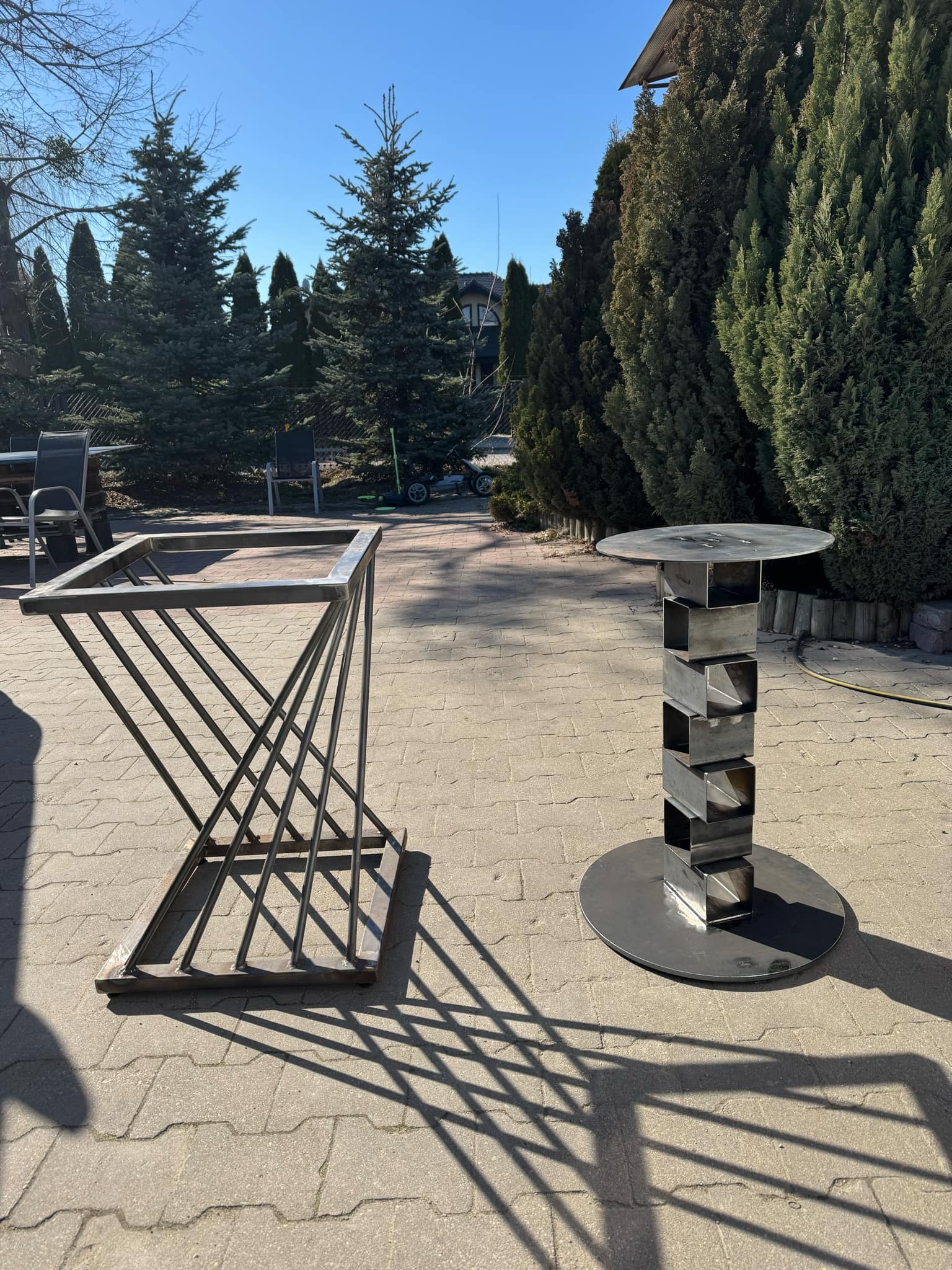 Dwie metalowe konstrukcje: geometryczna podstawa i stolik z kwadratowych elementów, na tle brukowanej powierzchni i zieleni. Słoneczny dzień.
