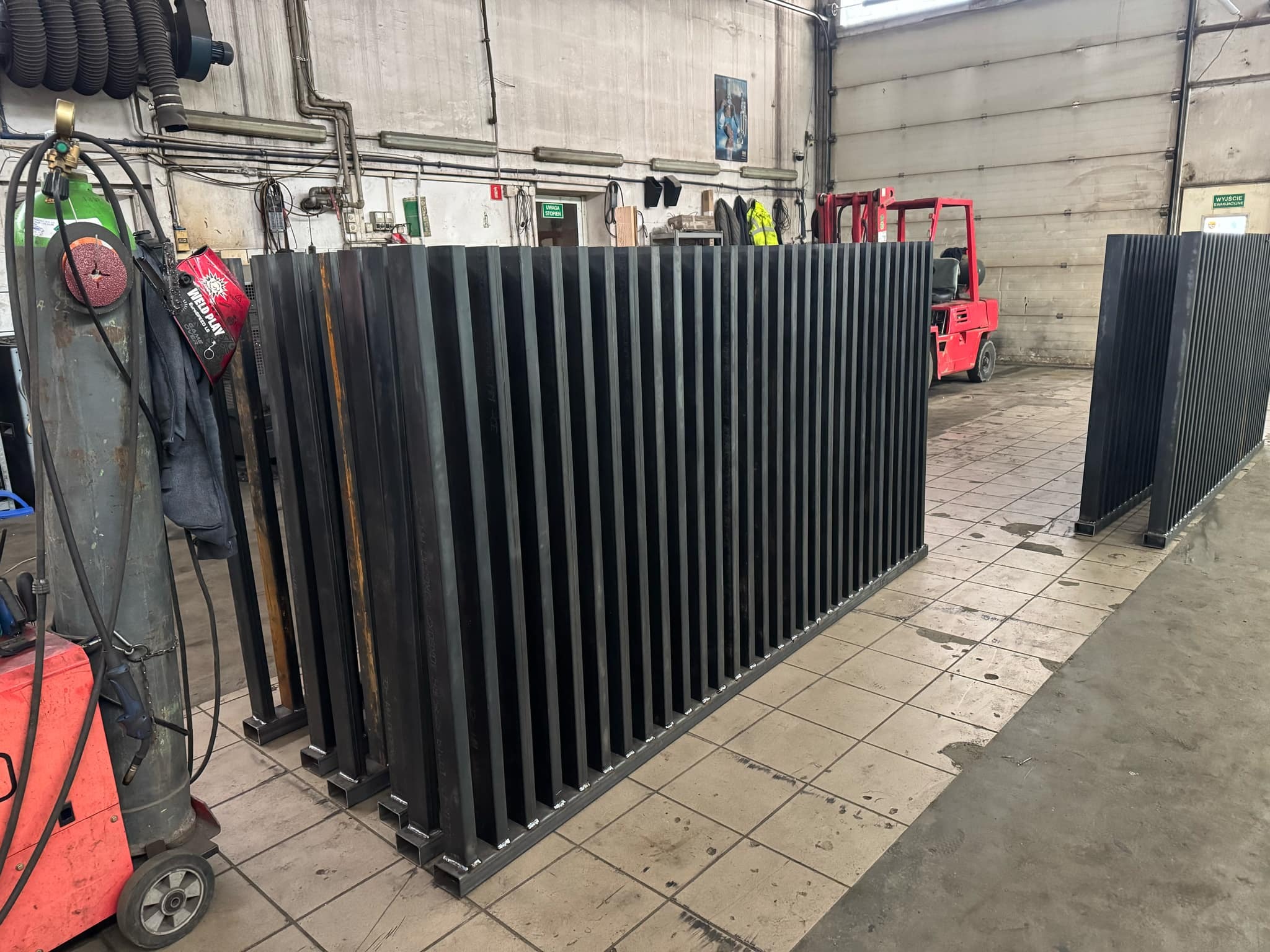 Czarne, metalowe elementy konstrukcyjne z pionowymi prętami, ustawione w hali produkcyjnej obok spawarki i wózka widłowego. Gotowe do montażu.