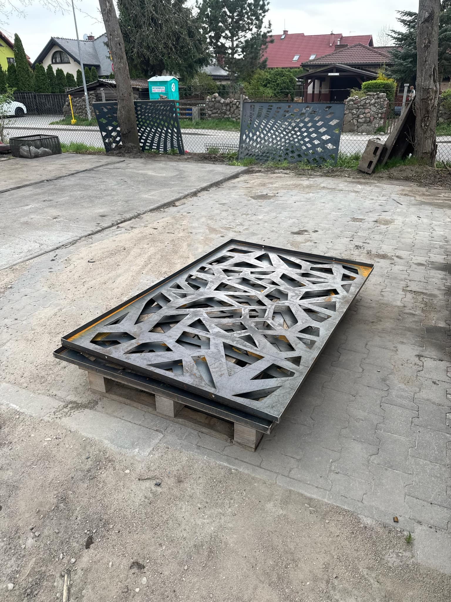 Metalowy panel dekoracyjny z ażurowym wzorem geometrycznym, ułożony na palecie na zewnątrz, prawdopodobnie element ogrodzenia lub balustrady.