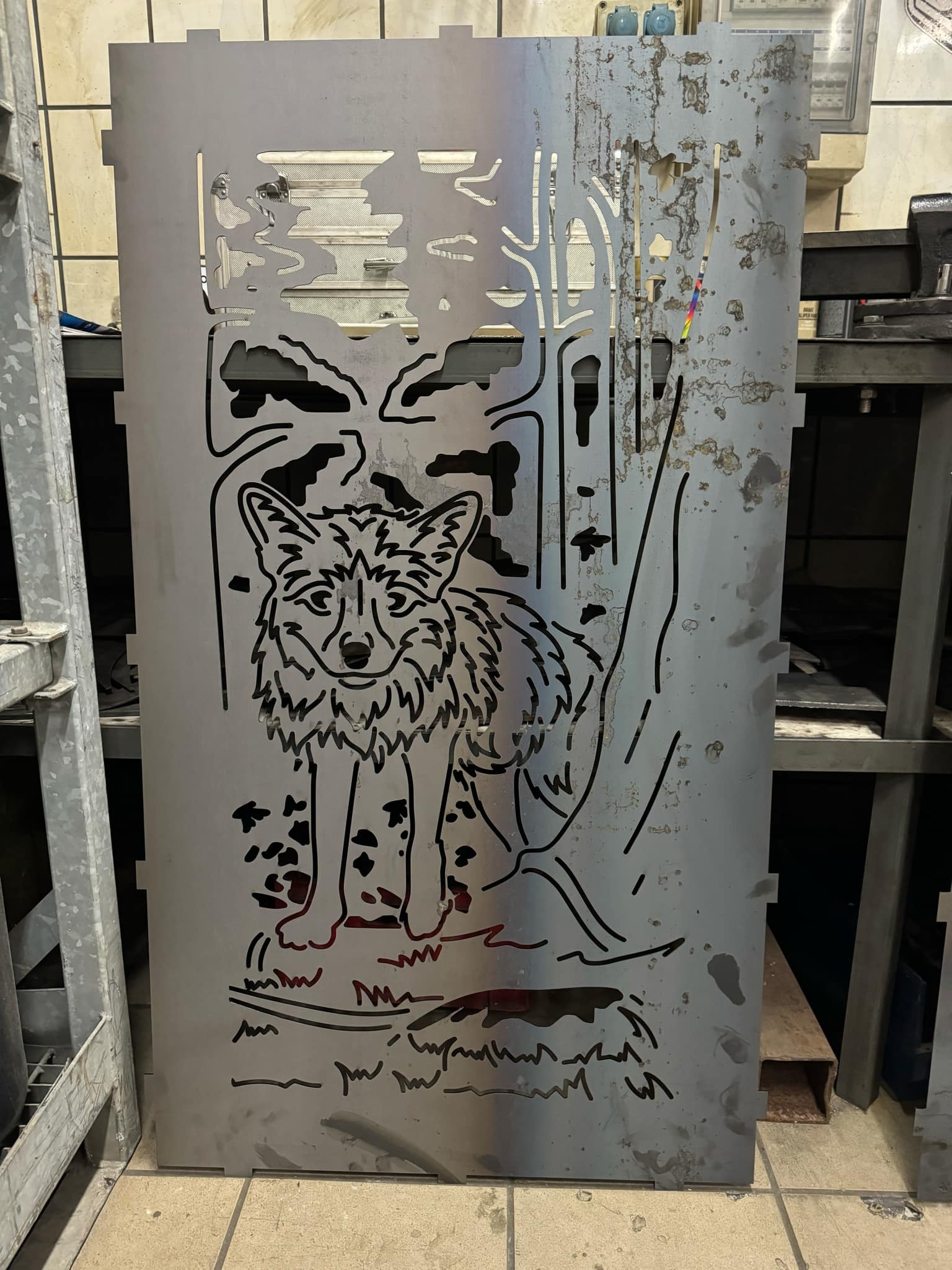 Metalowy panel z wyciętym motywem wilka na tle lasu, wykonany techniką CNC, oparty o metalowy regał. Widoczne ślady obróbki i zabrudzenia.