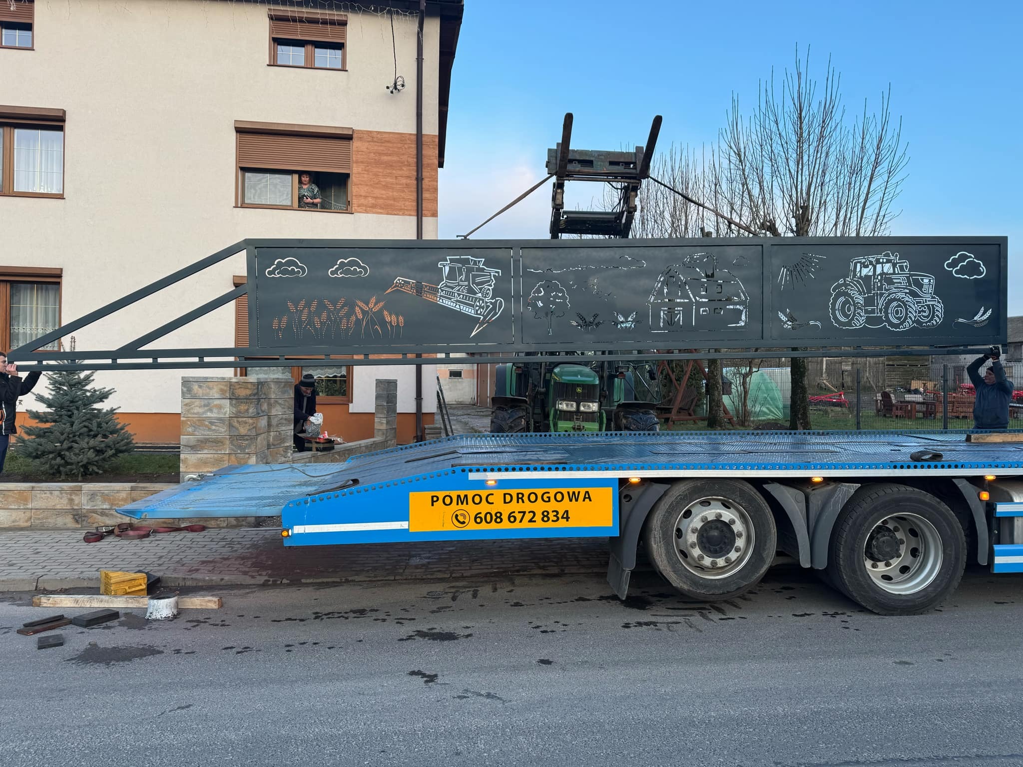 Transport bramy z wycinanymi laserowo wzorami wiejskimi na lawecie pomocy drogowej. W tle dom z oknem, w którym stoi osoba. Widoczny traktor i fragmenty montażu.