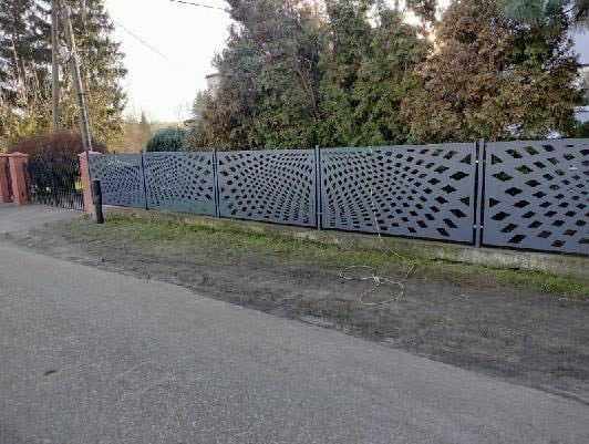 Nowoczesne ogrodzenie panelowe z geometrycznym wzorem, szare panele z wycinanymi rombami, widok z perspektywy ulicy, zieleń w tle.