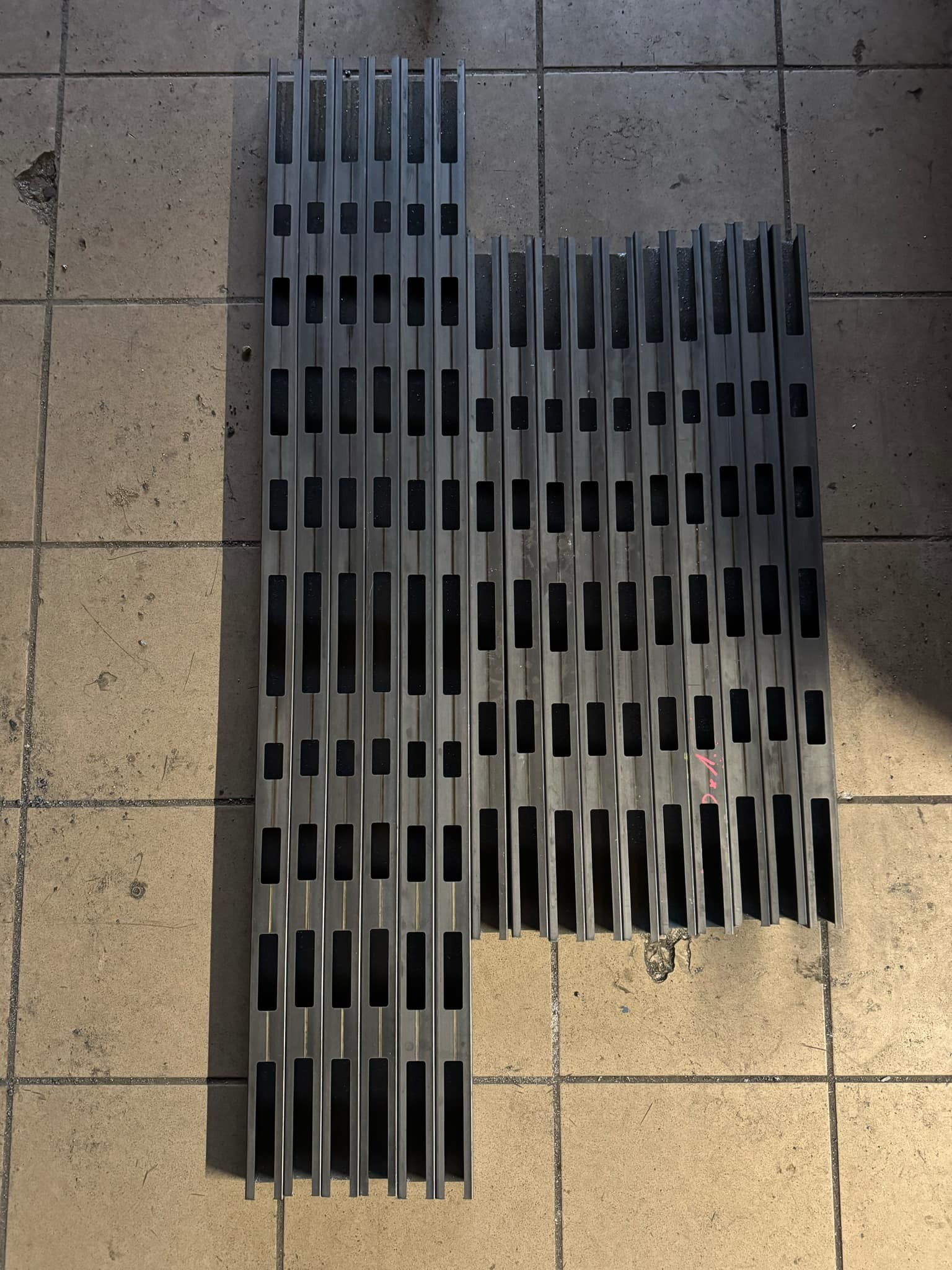 Metalowe profile z prostokątnymi otworami ułożone na podłodze z płytek. Elementy konstrukcyjne do balustrad lub ogrodzeń, widoczne ślady obróbki.