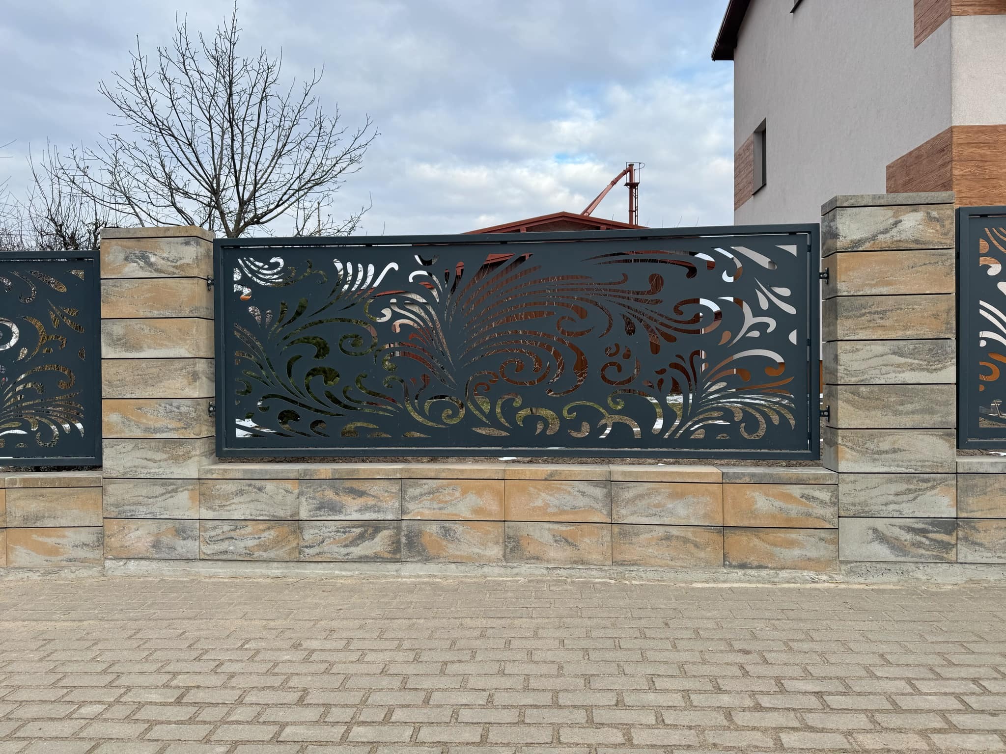 Dekoracyjna, metalowa brama wjazdowa z laserowo wycinanym wzorem roślinnym, zamontowana na słupkach z kamiennych bloków. W tle budynek i fragment żurawia.