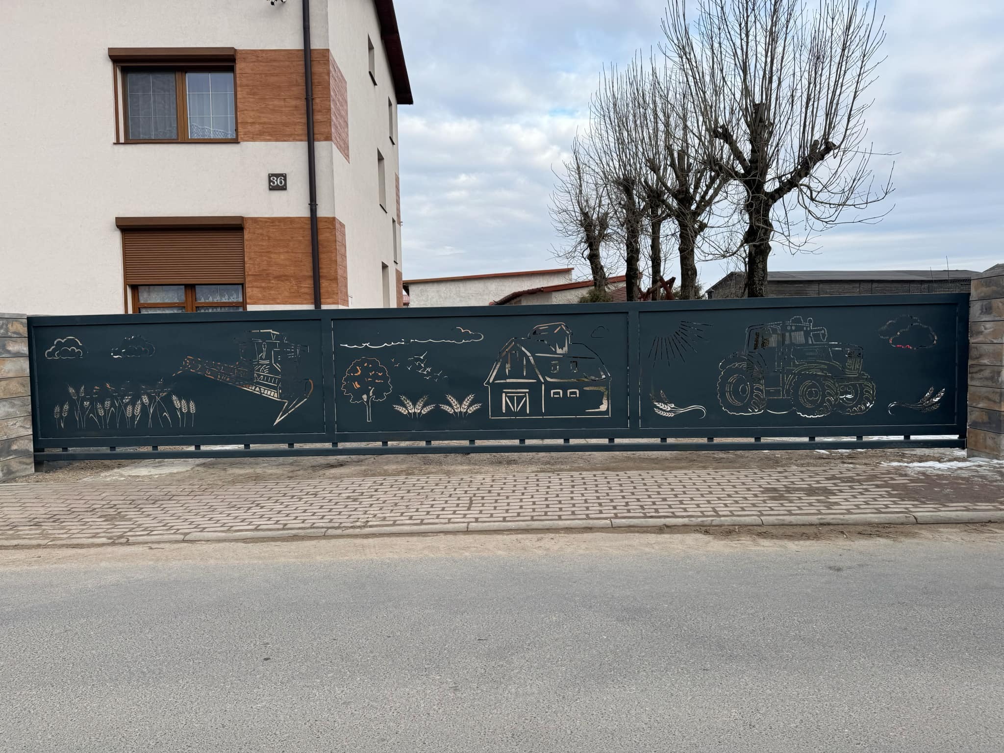 Metalowa brama przesuwna z motywami wiejskimi (traktor, zboże, dom) w Iławie. Ciemna brama na tle jasnego budynku i szarej drogi. Unikalne, ażurowe wzory.