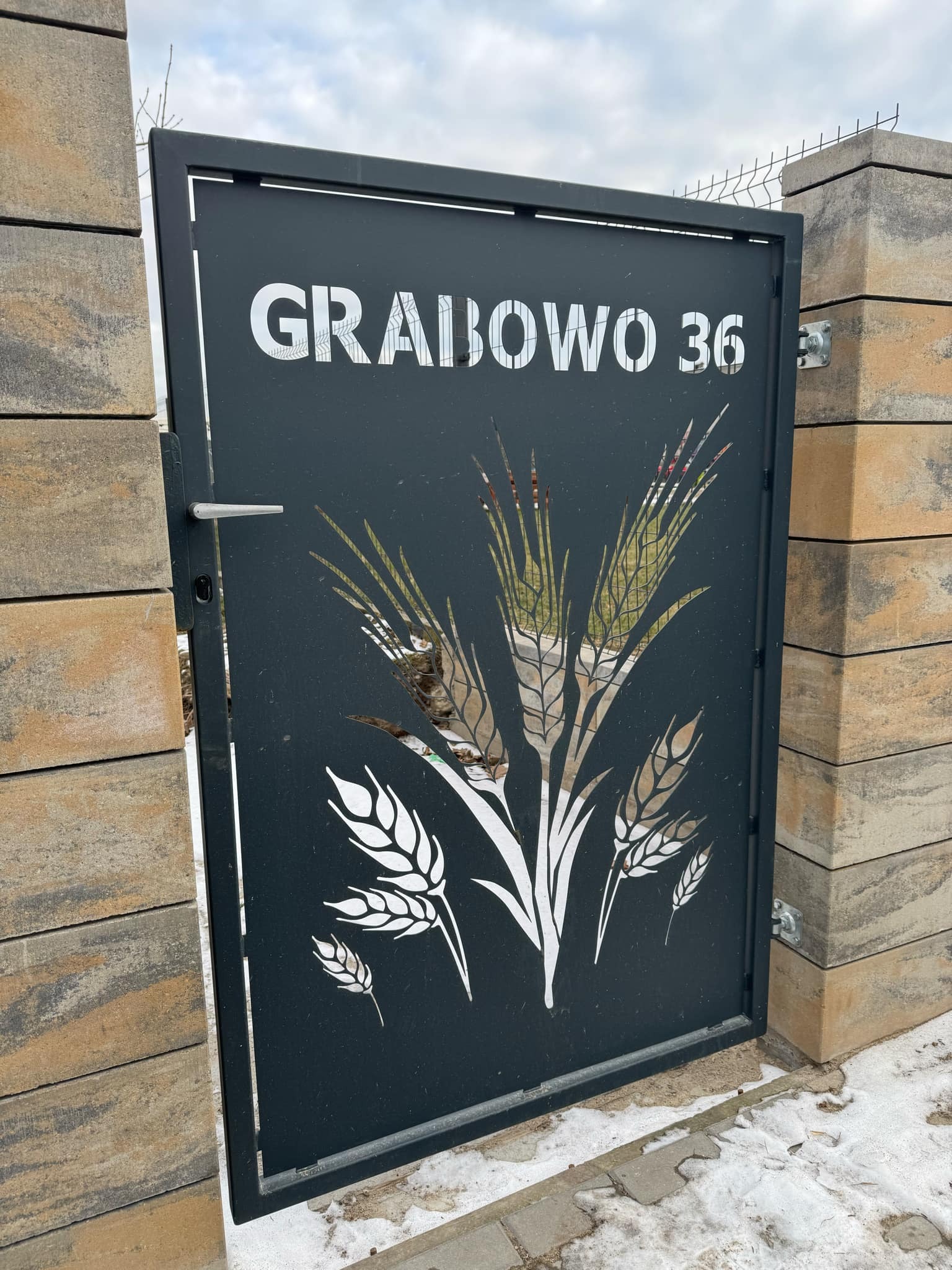 Nowoczesna brama wjazdowa z wycinanym laserowo wzorem kłosów zboża i numerem domu 'Grabowo 36', zamontowana na słupkach z bloczków betonowych.