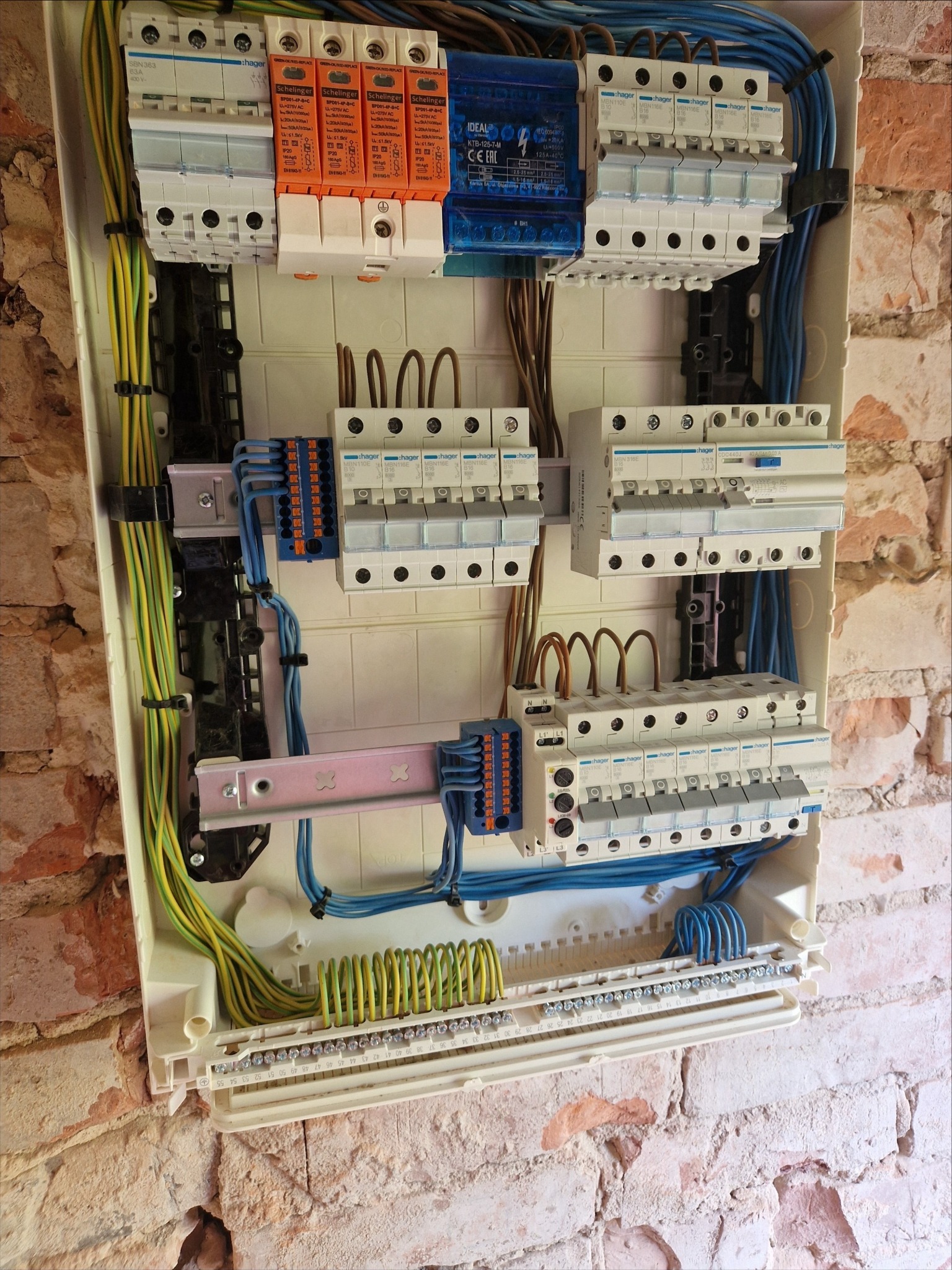 Rozdzielnia elektryczna Hager z bezpiecznikami i przewodami na tle ceglanej ściany, widok z bliska. Instalacja schludna, kable uporządkowane, widoczne oznaczenia.