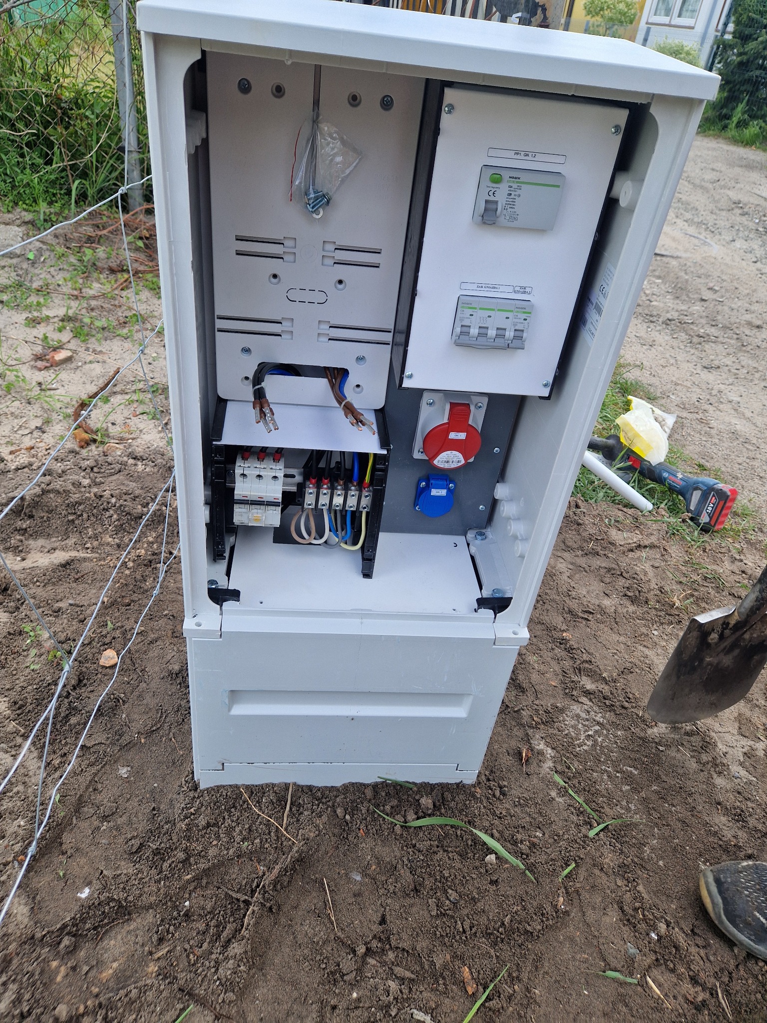 Otwarta, biała skrzynka elektryczna z widocznymi bezpiecznikami, gniazdami i przewodami. Obok leży wiertarka i szpadel, sugerujące prace instalacyjne w toku.