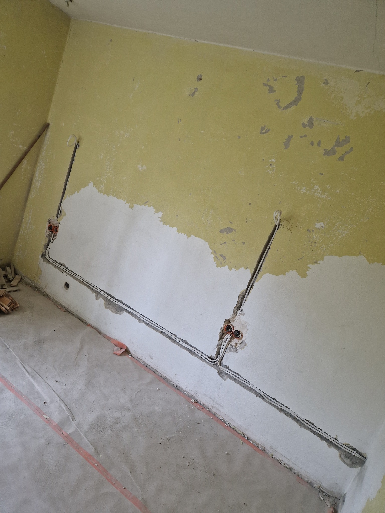 Wnętrze w trakcie remontu: żółta ściana z częściowo położoną gładzią, widoczne kable elektryczne w bruzdach i puszki instalacyjne. Podłoga zabezpieczona folią malarską.