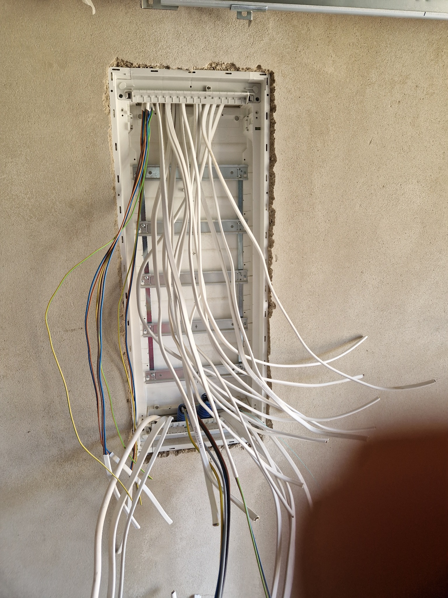 Rozdzielnica elektryczna w ścianie, z widocznymi przewodami w różnych kolorach. Surowa ściana w tle. Instalacja w trakcie montażu, widoczne elementy mocujące.