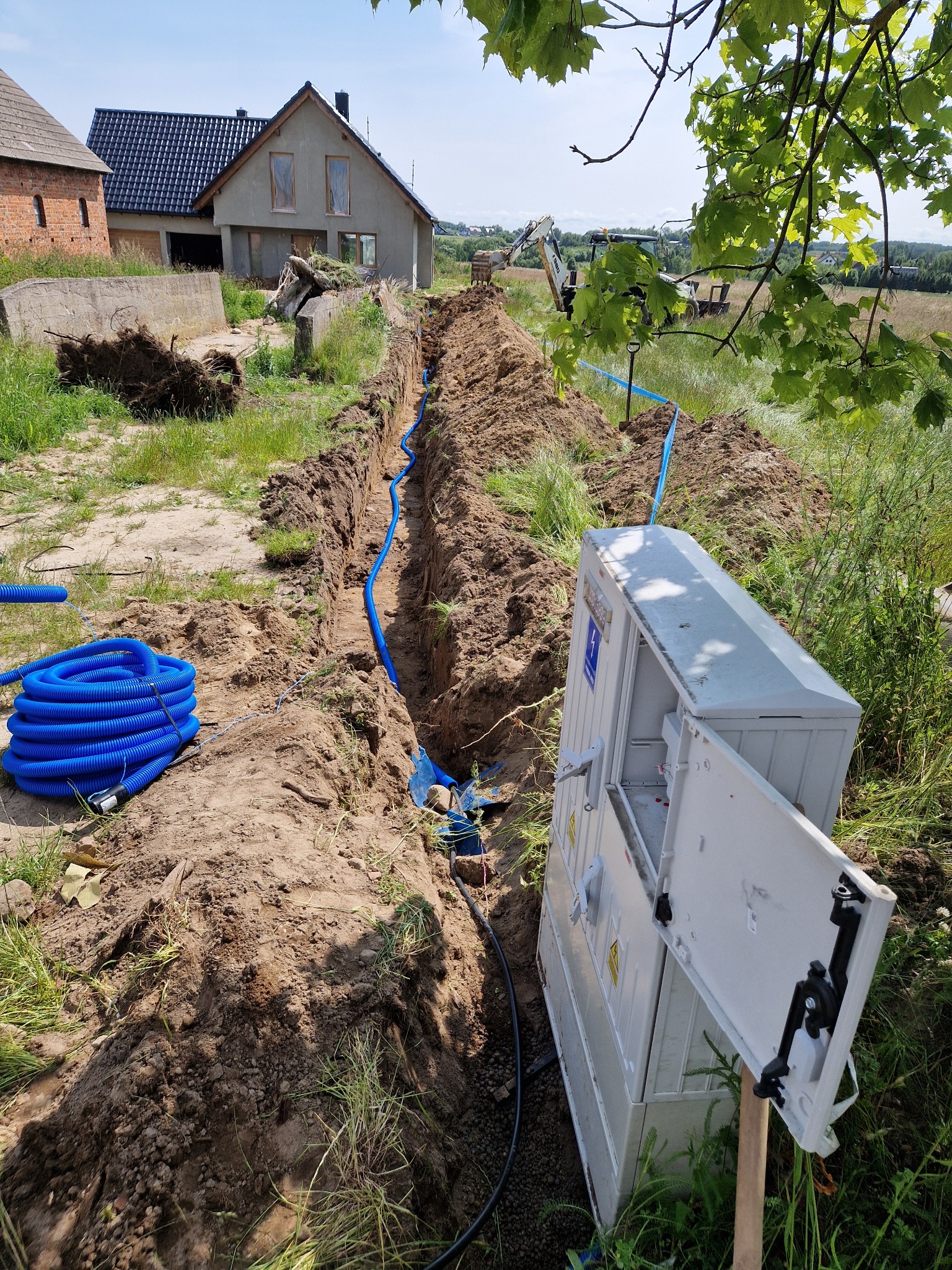 Głęboki wykop w ziemi z niebieskim przewodem, otwartą skrzynką rozdzielczą i zrolowanym kablem. W tle dom w budowie i koparka.