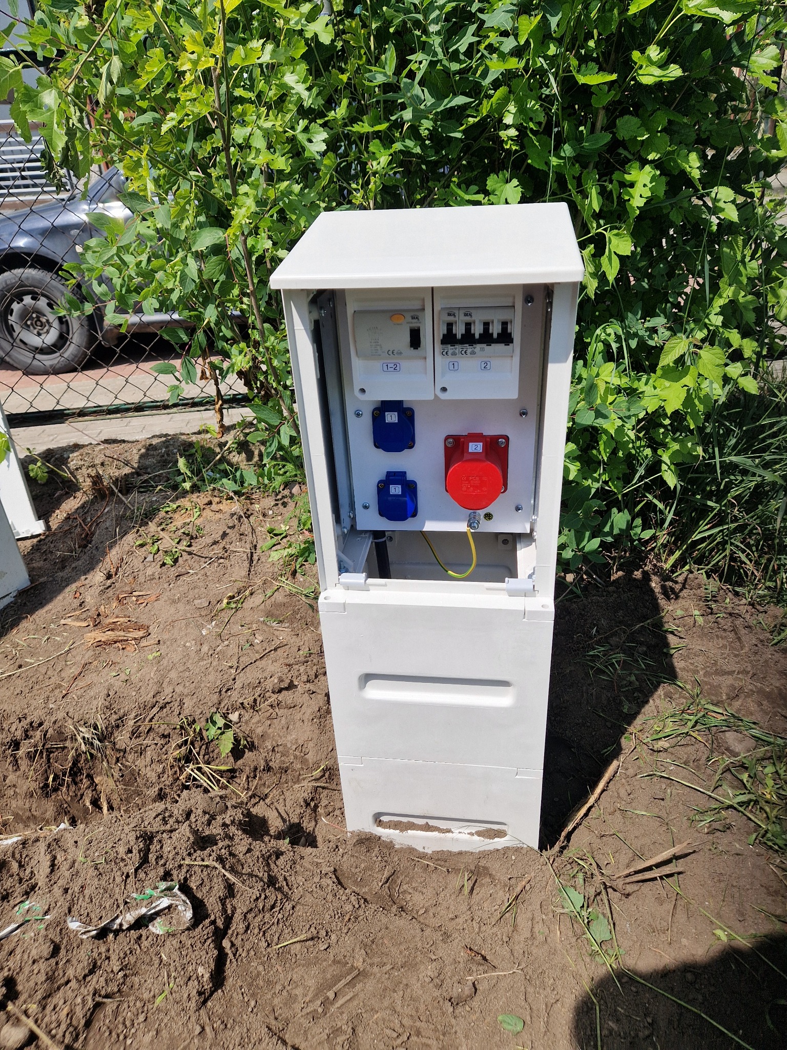 Biała skrzynka elektryczna na zewnątrz z gniazdami i wyłącznikami, umieszczona w ziemi, otoczona zielenią. Widoczne gniazda w kolorze niebieskim i czerwonym.