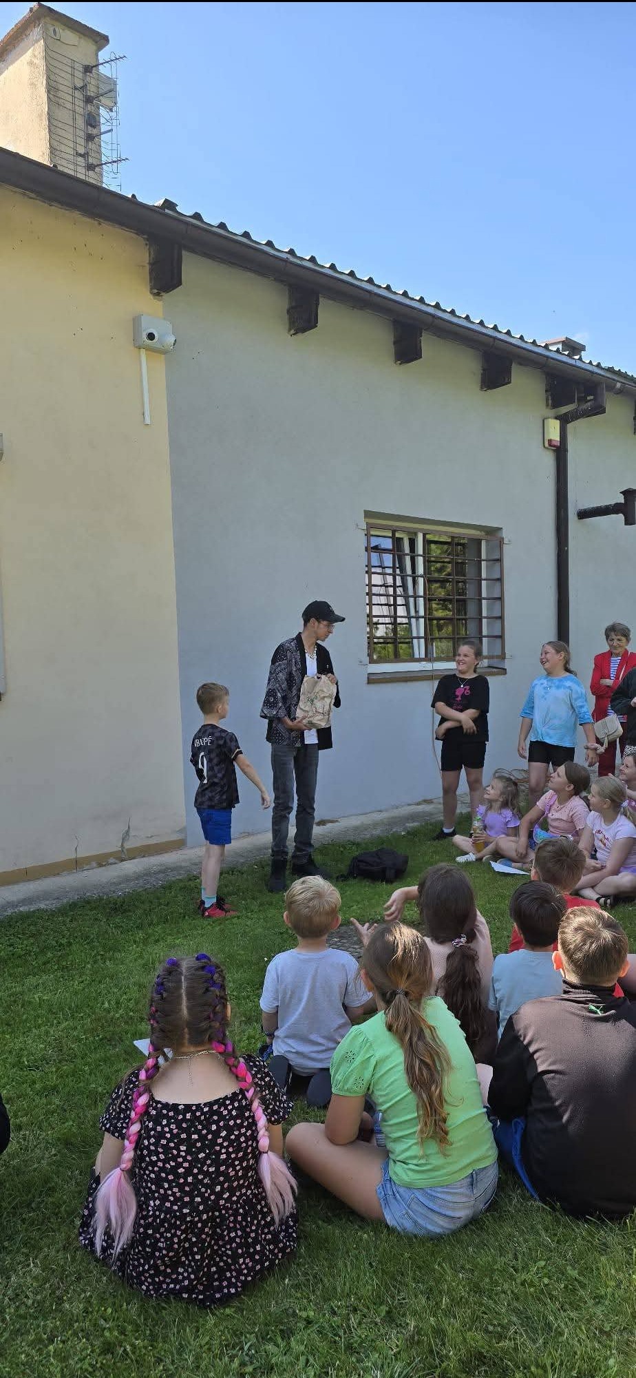 Animator w czapce i wzorzystej koszuli prezentuje rekwizyt przed grupą dzieci siedzących na trawie przed budynkiem podczas imprezy plenerowej.