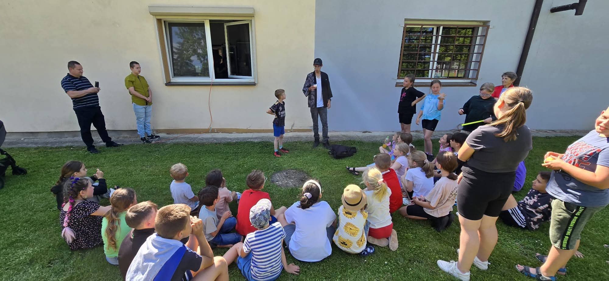 Animator w trakcie występu dla dzieci siedzących na trawie przed budynkiem. Widoczni dorośli obserwujący. Dzień słoneczny, jasne oświetlenie.