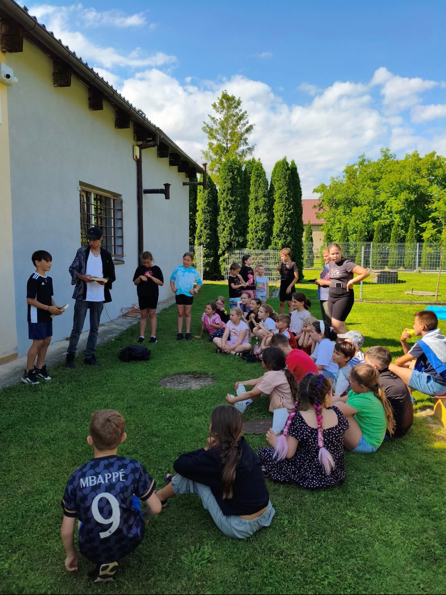 Animatorzy prowadzą zajęcia na trawie dla grupy dzieci, prezentując książki. Uczestnicy siedzą w kręgu, budynek w tle, słoneczny dzień.